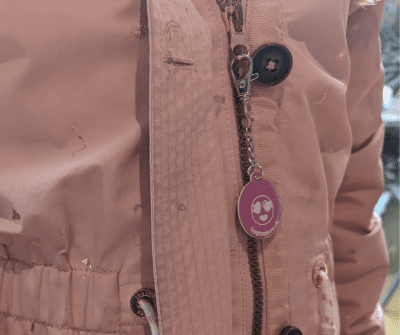 Image is zoomed in to show a girls pink coat with wooden buttons and a zip - attached to the zip is a pink circle tinytag with heart eyes and smile connected to the zip a with a keychain
